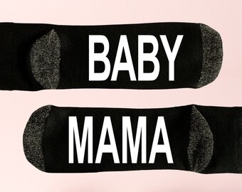 May include: Two black socks with white text. The top sock says "BABY" and the bottom sock says "MAMA". The socks are on a pink background with white flowers.