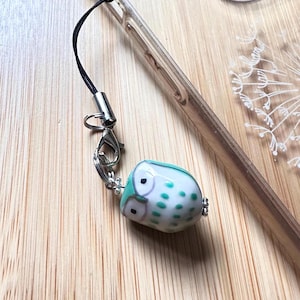 May include: A teal and white owl charm with black eyes. The charm is attached to a black phone strap with a silver clasp.
