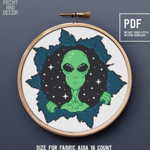 May include: A cross-stitch pattern featuring a green alien peeking out of a hole in a black and blue starry sky. The pattern is framed in a wooden hoop and is recommended for use with 16 count Aida fabric. The pattern is 131 stitches wide by 133 stitches high and requires 9 colors. The pattern is available as a PDF download.