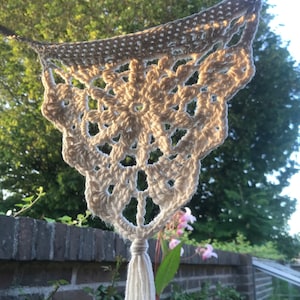 Puede incluir: Un tapiz de macramé blanco tejido a crochet con un borla. El tapiz tiene forma de triángulo con un diseño floral en el centro.