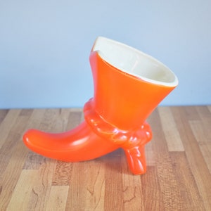 May include: An orange ceramic boot-shaped vase with a white interior. The vase has a knotted detail around the ankle and a small heel. The boot is set against a light blue background and a wooden surface.