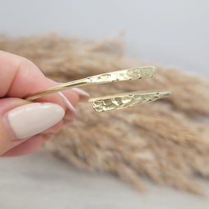 May include: A gold hammered metal bangle bracelet with a simple, open design. The bracelet is made of a single piece of metal that is bent into a circle and then hammered to create a textured surface.