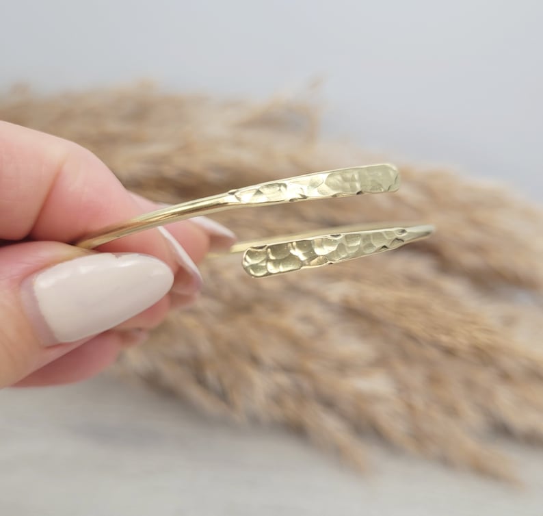 May include: A gold hammered metal bangle bracelet with a simple, open design. The bracelet is made of a single piece of metal that is bent into a circle and then hammered to create a textured surface.