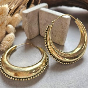 May include: A pair of gold-toned hoop earrings featuring a thick, crescent-shaped design with a polished finish. The outer edges of each hoop are detailed with a delicate row of small, raised beads, and each earring includes a thin wire post closure.