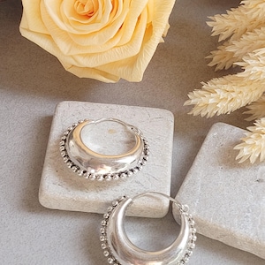 May include: A pair of silver hoop earrings with a crescent shape. The earrings have a smooth, polished inner surface and a beaded border. The earrings are displayed on a gray surface with a yellow rose and dried flowers in the background.