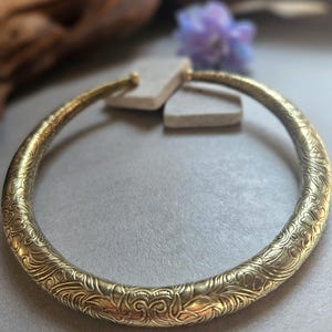 May include: A gold-toned, patterned collar necklace. The necklace has intricate floral and swirl designs across its surface. The design is a rigid, circular shape, and the metal has a polished finish. The background is blurred, with hints of wood and a purple flower.