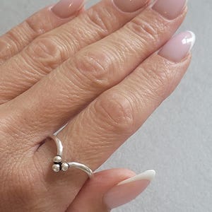 May include: A silver ring with a V-shaped design, featuring three small, round embellishments. The ring is worn on a finger with a French manicure, showcasing pale pink nails with white tips.