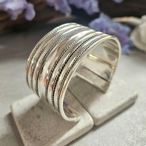 Op de afbeelding: Een brede, zilverkleurige manchetarmband. De armband heeft meerdere parallelle banden met een getextureerd, touwachtig ontwerp tussen elke band. De manchet wordt weergegeven op een lichtgrijze ondergrond, met vage paarse bloemen op de achtergrond.