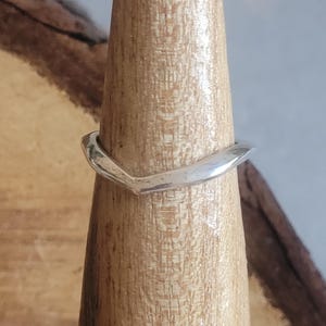 May include: A silver ring with a V-shaped design. The ring is displayed on a wooden ring holder. The ring's surface has a slightly textured appearance, reflecting light. The ring is a piece of jewellery.