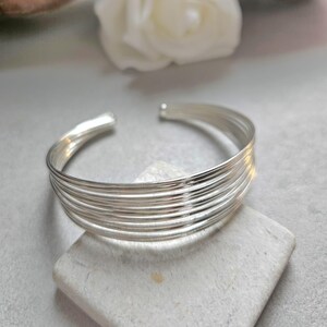 Può includere: Un bracciale a polsino argentato con più bande parallele. Il design aperto consente di indossarlo facilmente. Il bracciale è esposto su una piccola pietra colorata, con un fiore bianco sfocato sullo sfondo.