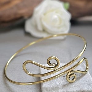 May include: A gold-colored arm cuff bracelet with a decorative swirl design. The bracelet features two spiral accents and a curved band. The jewelry is displayed on a light gray surface with a blurred white flower in the background.