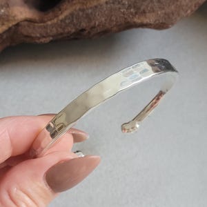 Może przedstawiać: Srebrna bransoletka typu cuff z młotkowaną fakturą. Otwarta konstrukcja ma zaokrąglony koniec. Bransoletka jest prezentowana na neutralnym tle, podkreślając jej prostą elegancję. Obwód ok. 17 cm.