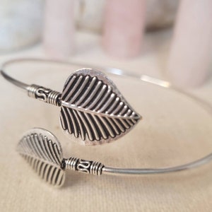 May include: Silver bangle bracelet with two leaf-shaped charms. The bracelet is thin and delicate, and the leaves are detailed with lines.