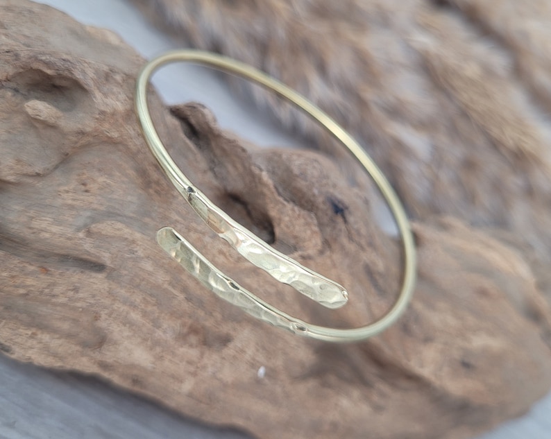 May include: A gold hammered bangle bracelet with a unique open design. The bracelet is made of a single piece of metal that is bent into a circle and then hammered to create a textured surface.
