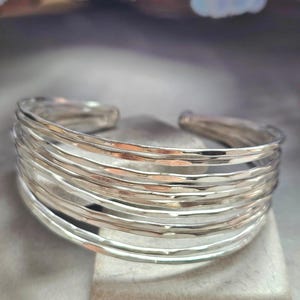 Op de afbeelding: Een zilveren manchetarmband met een gehamerde textuur. De open armband heeft meerdere parallelle banden, waardoor een gelaagd effect ontstaat. De sieraden worden gepresenteerd op een neutraal oppervlak, met zachte, vage achtergrondelementen.