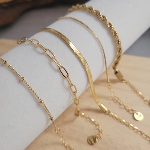Peut inclure: Quatre colliers dorés de différents modèles sont présentés. Ils comprennent une chaîne perlée, une chaîne à maillons ovales, une chaîne en chevrons et une chaîne torsadée. Chaque collier a une chaîne délicate et réglable avec un petit charme rond.
