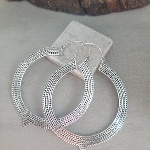 May include: A pair of large, silver hoop earrings. The earrings feature a detailed design with multiple rows of small, raised dots and a triangular accent at the base. The hoops are displayed on a grey surface.