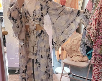 May include: A two-piece set with a tie-front top and a matching skirt. The set features a light blue and black tie-dye pattern. The top has wide sleeves. The skirt is long and flowing. The outfit is displayed in a boutique setting.
