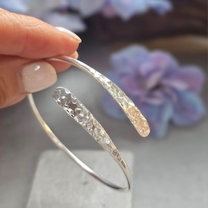 Puede incluir: Una pulsera de brazalete plateada con una textura martillada en los extremos. La pulsera tiene un diseño abierto, con los extremos curvándose ligeramente uno del otro. El fondo está borroso, con toques de morado y blanco.