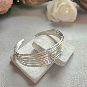 Op de afbeelding: Een zilveren manchetarmband met meerdere parallelle banden. De armband wordt getoond op een kleine, lichtgrijze rechthoekige steen. Een witte roos en andere decoratieve elementen zijn op de achtergrond.