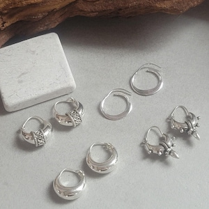 May include: A collection of silver earrings in various designs. The earrings include simple hoops, and others with intricate details like beaded or spiked accents. The jewelry is displayed on a light gray surface, showcasing the different styles.