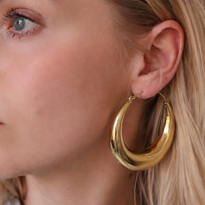 May include: Close-up of a person's face with a gold-colored hoop earring. The earring is a large, oval shape and reflects light. The person has blonde hair and fair skin.