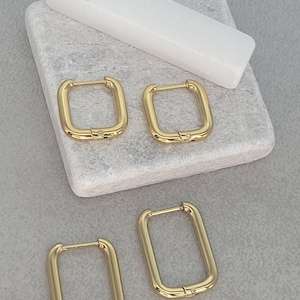 Op de afbeelding: Vier goudkleurige rechthoekige oorringen. De oorbellen worden gepresenteerd op een lichtgrijze stenen ondergrond met een wit rechthoekig object. De oorbellen hebben een eenvoudig, modern design.