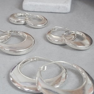 以下が含まれることがあります： グレーの表面に、いくつかのペアのシルバーフープピアスが展示されています。イヤリングは滑らかで磨かれた仕上げで、丸みを帯びた三日月型をしています。背景には、小さな白い長方形のオブジェクトがあります。