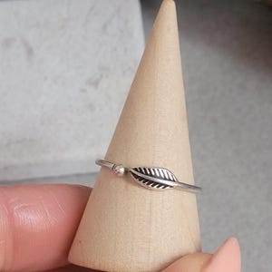 May include: A silver ring with a leaf design. The ring has a small, round bead and a detailed leaf shape. The ring is displayed on a wooden cone. The ring is a delicate piece of jewellery.