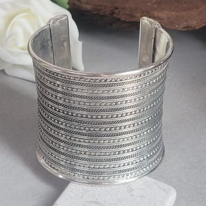 Può includere: Un ampio bracciale a polsino argentato con intricati dettagli incisi orizzontalmente. Il bracciale ha un'apertura rettangolare ed è esposto su una superficie grigio chiaro. Il design del bracciale presenta file di piccoli motivi quadrati e linee sottili e strutturate.