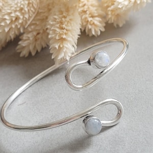 May include: A silver open cuff bracelet featuring two looped ends, each set with a small, round, translucent moonstone cabochon. The polished silver band curves into a bypass design, showcasing the iridescent white stones at each terminal point.