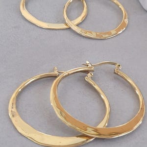 May include: Two pairs of gold-coloured hoop earrings. The earrings have a hammered texture on the lower half of the hoops. The earrings are displayed on a light grey surface. The earrings are a classic jewellery item.