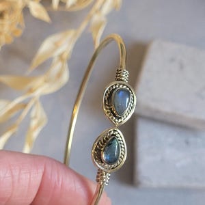 Puede incluir: Un brazalete de metal en tono dorado con dos piedras de labradorita en forma de lágrima. Cada piedra está engastada en un bisel decorativo de alambre trenzado, con detalles de metal enrollado que aseguran los engastes a la banda metálica fina.
