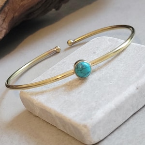 May include: A gold-colored open cuff bracelet with a round, turquoise-colored stone set in the center. The bracelet has a simple, elegant design and is displayed on a white stone surface.
