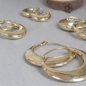 May include: A collection of gold-coloured hoop earrings, each with a smooth, polished surface and a crescent shape, are arranged on a light grey surface. The image is well-lit, showcasing the metallic sheen of the jewellery.