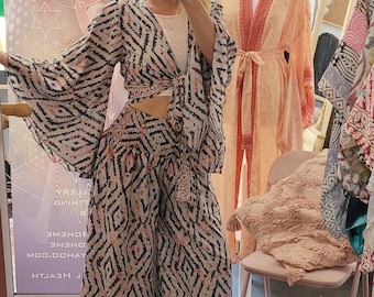May include: A two-piece bohemian outfit with a geometric pattern in black, white, and coral. The top is a wrap style with flared sleeves, paired with wide-leg pants. A peach-colored robe with coral trim is visible in the background.