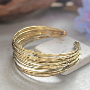 May include: A gold cuff bracelet with a layered, textured design. The bracelet is made of multiple strands of metal, creating a bold accessory. The open design allows for easy wear. A statement piece for any jewellery collection.