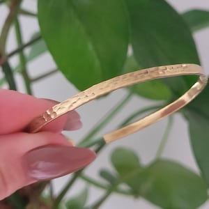 May include: A gold bangle bracelet with a hammered texture. The bracelet has a simple, open design, and the metal has a bright, polished finish. The background includes green leaves.