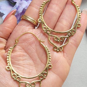 May include: A pair of gold-colored hoop earrings. The earrings have a detailed design with small beaded accents along the top and decorative filigree elements. The earrings are shown against a soft background.