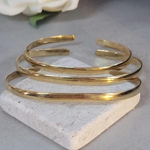 Può includere: Quattro braccialetti dorati aperti sono impilati su una piccola pietra quadrata bianca. I braccialetti hanno un design semplice e minimalista e una finitura lucida. Un fiore bianco è visibile sullo sfondo.