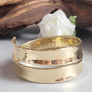 May include: A gold-coloured cuff bracelet with a hammered texture. The bracelet has a spiral design. A white rose and a piece of wood are in the background.