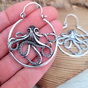 May include: Silver hoop earrings featuring an octopus design. The octopus is detailed with textured tentacles and a circular body.