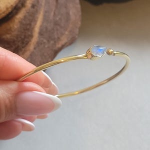 May include: A delicate gold-toned bracelet featuring a teardrop-shaped, iridescent moonstone. The bracelet has a simple, elegant design with a small gold bead accent. The jewelry is a beautiful accessory.