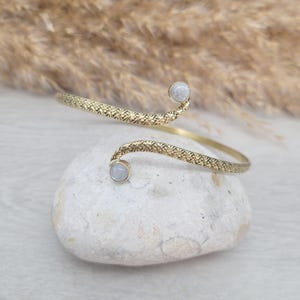 May include: A gold-coloured arm cuff with a textured pattern. The cuff has two curved ends, each with a small, round, pale-coloured stone. The cuff is on a white stone, with dried plants in the background.