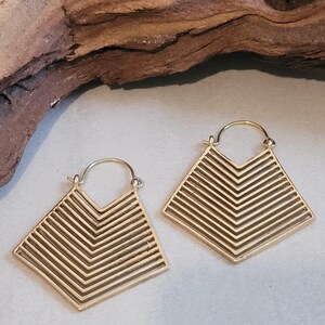 Pode incluir: Um par de brincos geométricos dourados. Cada brinco apresenta uma forma de losango com um design de galão feito de linhas paralelas. Os brincos têm um fecho de argola simples.