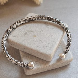 May include: Silver cuff bracelet with a geometric pattern. The open-ended bracelet has a repeating design of small, interconnected shapes. The ends of the bracelet have rounded, ball-shaped accents. The bracelet is displayed on a grey stone surface.
