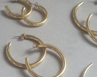 May include: Several pairs of gold-colored hoop earrings. The earrings are a simple, classic design with a smooth, rounded surface. The earrings are displayed on a light gray surface.