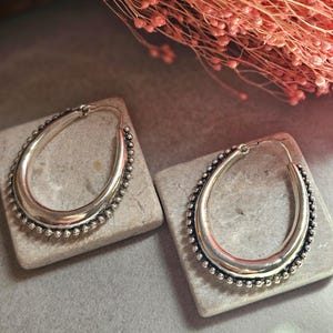 May include: A pair of silver-toned oval hoop earrings. The earrings have a smooth, polished inner surface and are bordered with small, round beads. The earrings are displayed on small, gray stones, with a blurred pink floral arrangement in the background.