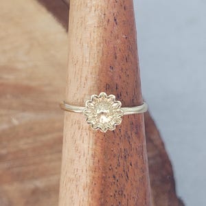 Può includere: Un delicato anello color oro con un design a forma di girasole. La fascia dell'anello è sottile e il girasole ha petali dettagliati e un centro strutturato. L'anello è esposto su un porta anelli di legno.
