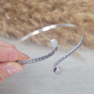 Pode incluir: Uma pulseira de punho prateada com um design texturizado. A pulseira apresenta duas pequenas pedras redondas e claras. O design aberto permite um uso fácil. É uma peça de joalharia.